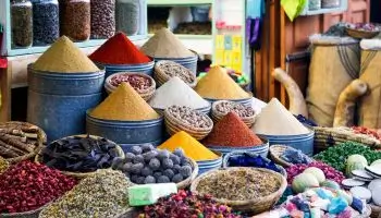 Zdjęcie przedstawia barwne stoisko targowe, najprawdopodobniej na orientalnym bazarze, z szerokim wyborem przypraw i ziół eksponowanych w efektowny, artystyczny sposób. W centralnej części widoczne są wysokie, stożkowate kopce przypraw usypane w metalowych naczyniach – mają intensywne kolory: czerwony (np. papryka lub chili), pomarańczowy (kurkuma), żółty, biały, beżowy, brązowy i ciemnobrązowy. Każda przyprawa uformowana jest w precyzyjny stożek, co jest charakterystyczne dla tradycyjnych targów w krajach Bliskiego Wschodu i Afryki Północnej. Niżej i na pierwszym planie znajdują się kosze i misy wypełnione suszonymi płatkami kwiatów, mieszankami ziół, kulkami zapachowymi i innymi aromatycznymi produktami. Wśród nich można dostrzec m.in. suszoną lawendę, hibiskus, czarne kule (prawdopodobnie suszone limonki), a także mydła i barwniki. Tło przedstawia sklepik z półkami wypełnionymi dodatkowymi produktami w workach i pojemnikach. Całość tętni kolorami, fakturami i zapachem, oddając autentyczny klimat lokalnego rynku przypraw.